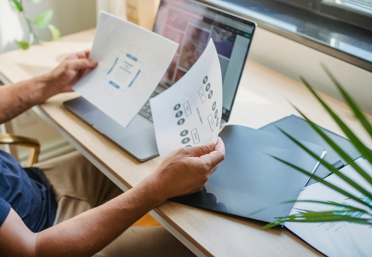 a freelance web designer showing two arms holding papers i front of a laptop on desk